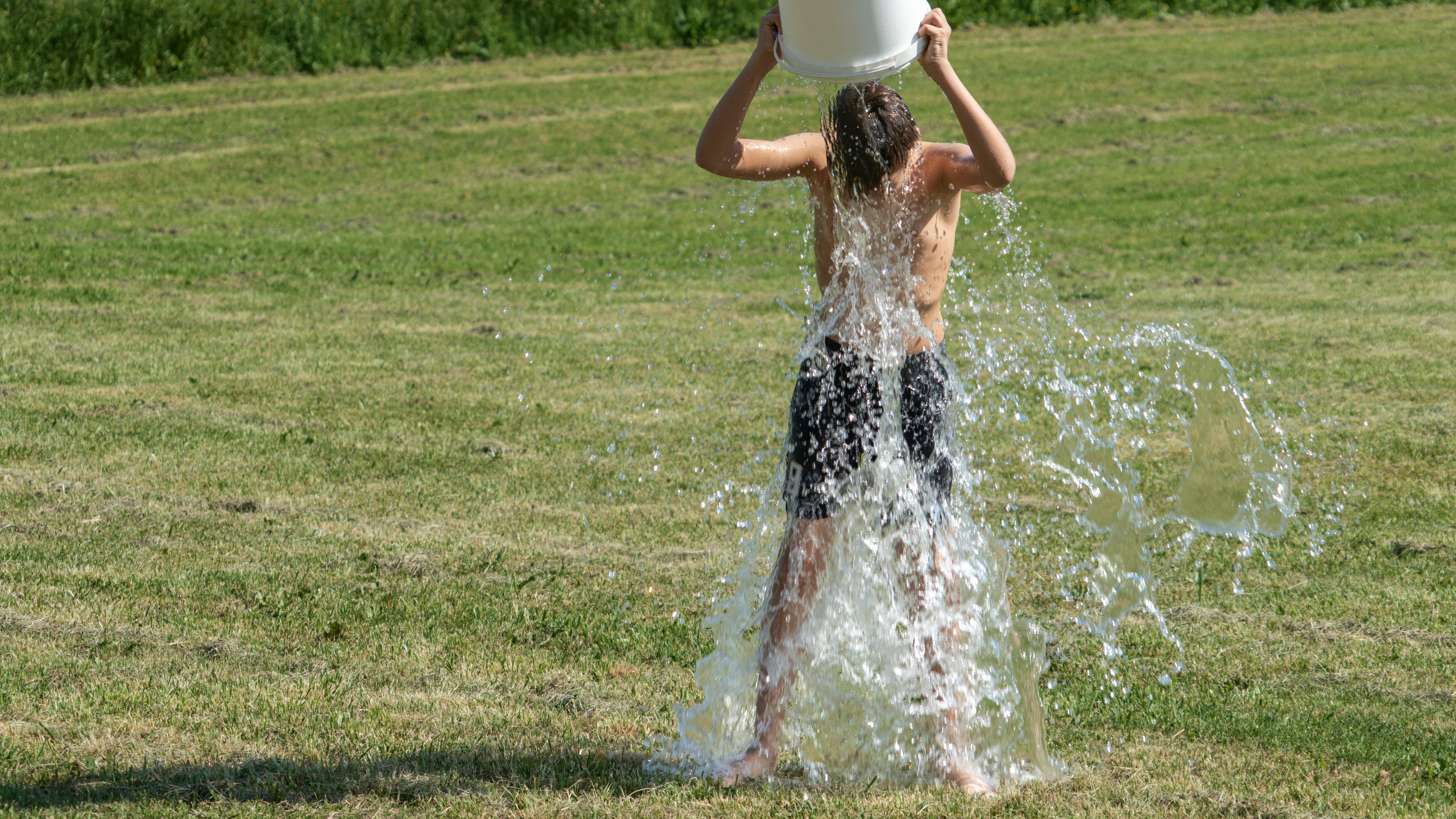 Teenage boy pouring bucket of cold water over his head outdoors.